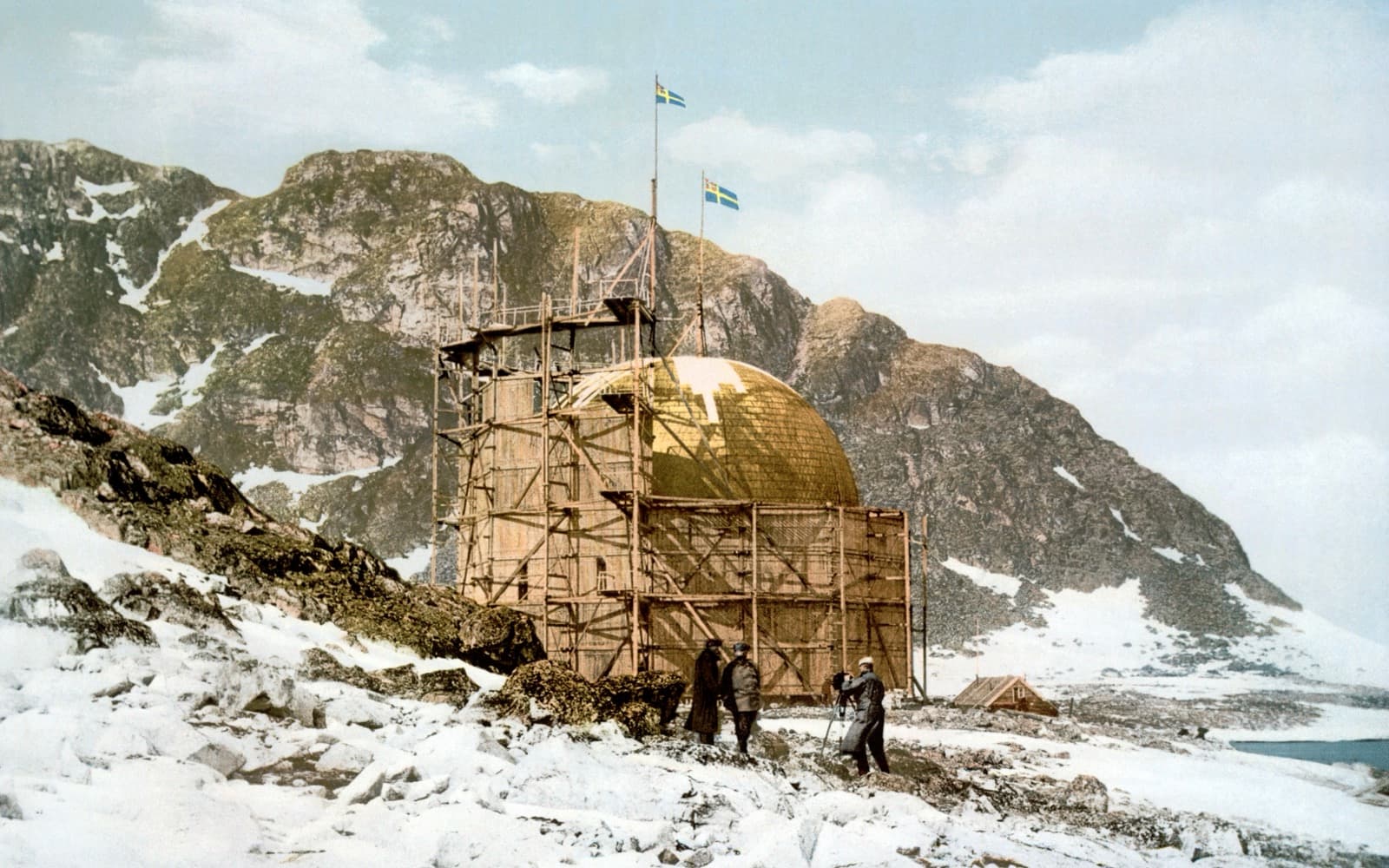 Andrées ballonghus av trä med två svenska flaggor framför ett snötäckt berg på Danskön.