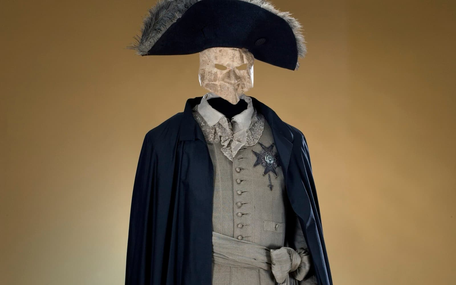 En maskeraddräkt med trekantig hatt, vit mask, svart mantel och grå väst med en ordensstjärna