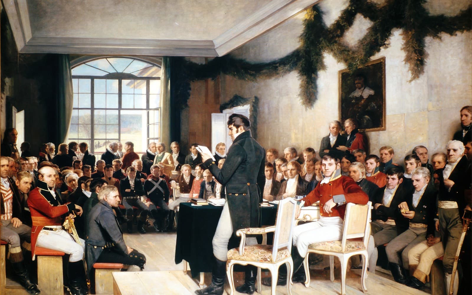 Målning av riksförsamlingen på Eidsvoll 1814, där en man står och läser upp ett papper inför en sal full av män i uniform och frack.