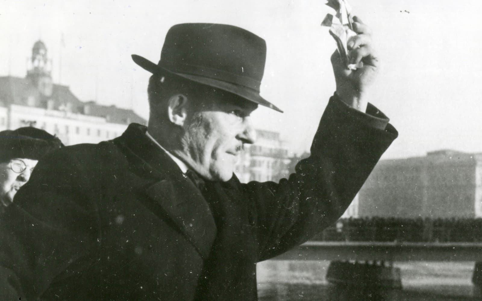 Svartvitt foto av en man i hatt och rock som håller upp en motbok vid en kaj
