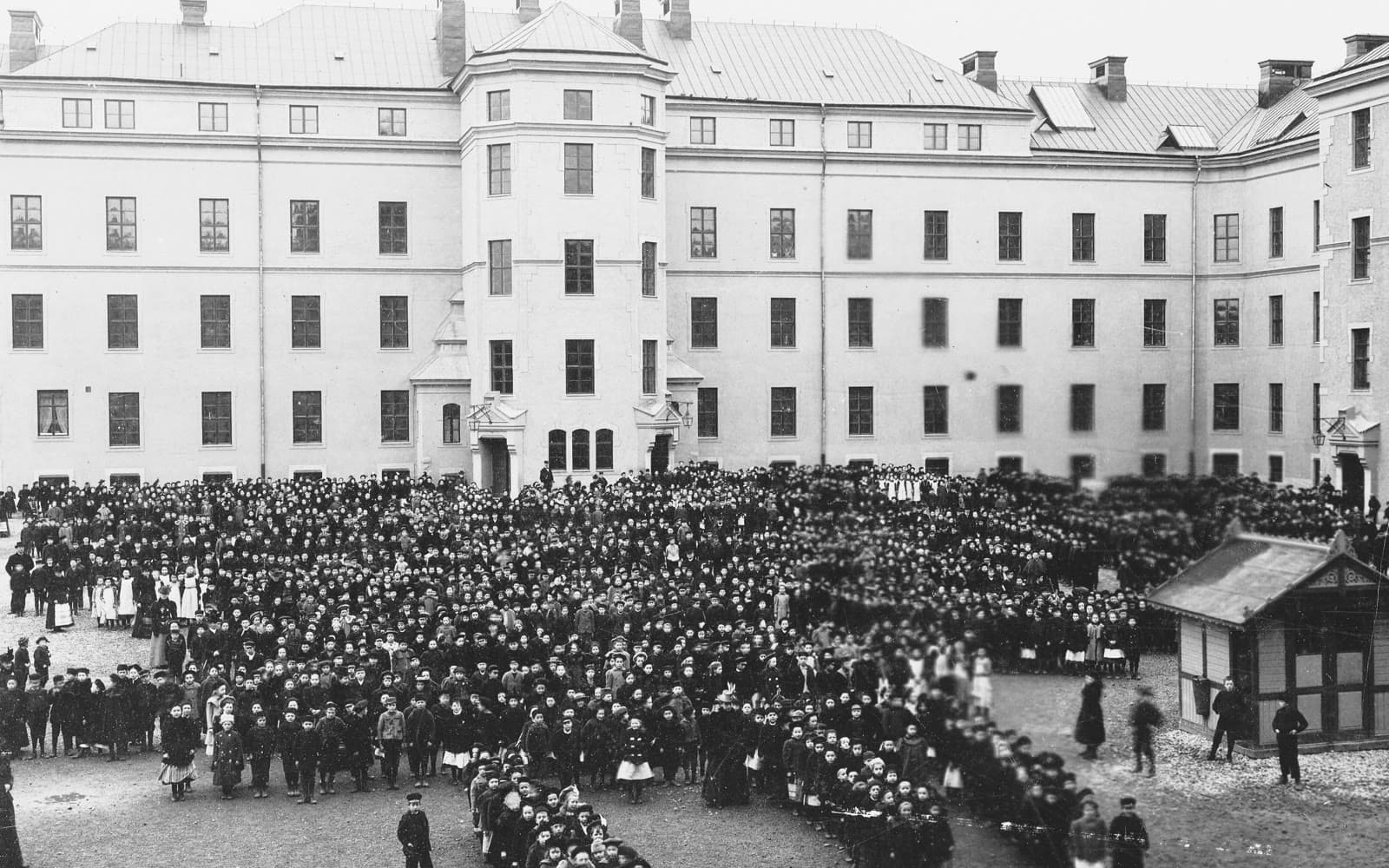 Svartvitt foto av en stor skolgård full av barn framför en hög skolbyggnad i Stockholm.