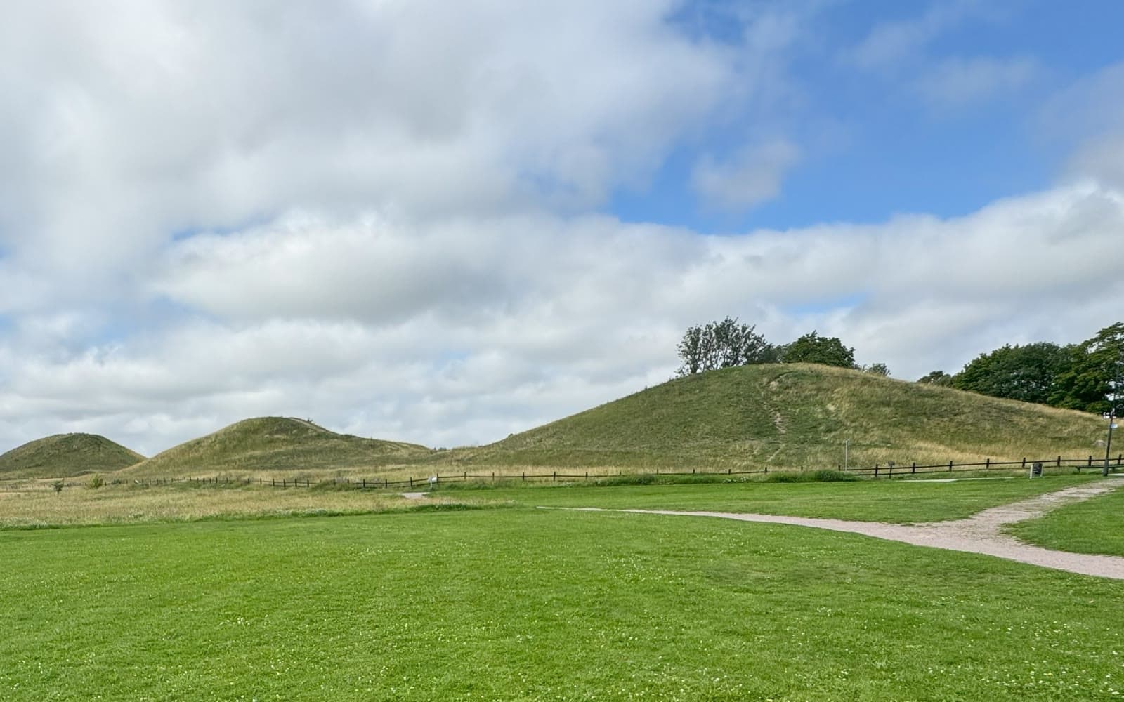 Tre stora gräsbevuxna gravhögar under en blå himmel med vita moln i Gamla Uppsala.