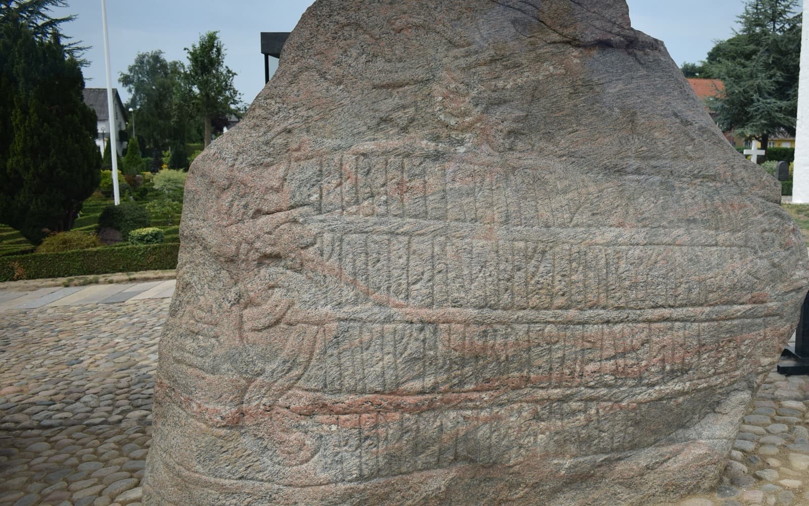 En stor grå runsten med inristade runor och slingor, på en stenlagd plats med träd i bakgrunden