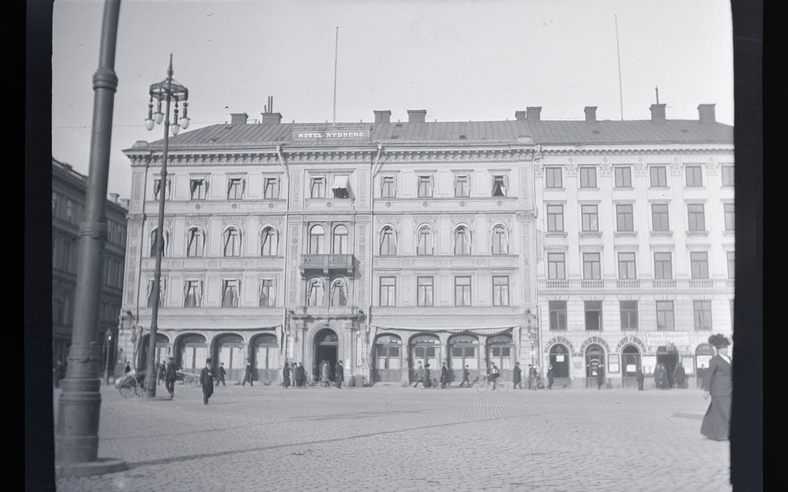 Fasaden på Hotel Rydberg vid ett kullerstensbelagt torg med människor och en gatlykta