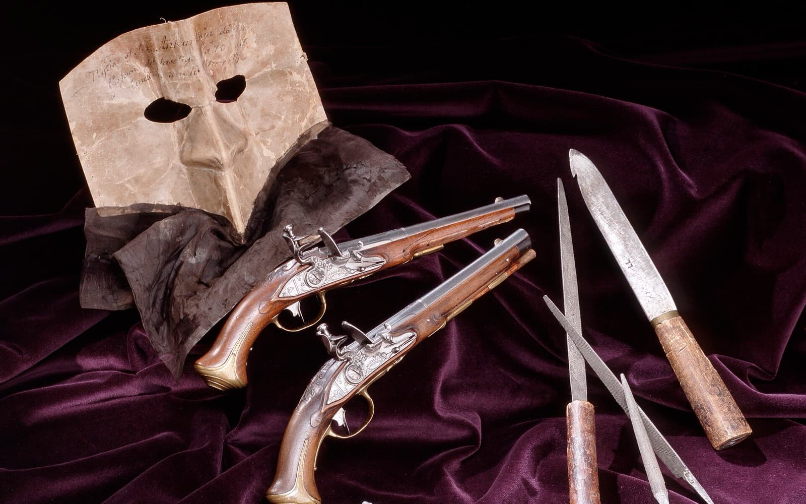 En vit mask, två flintlåspistoler och en kniv med blad och några små kulor på mörklila sammet