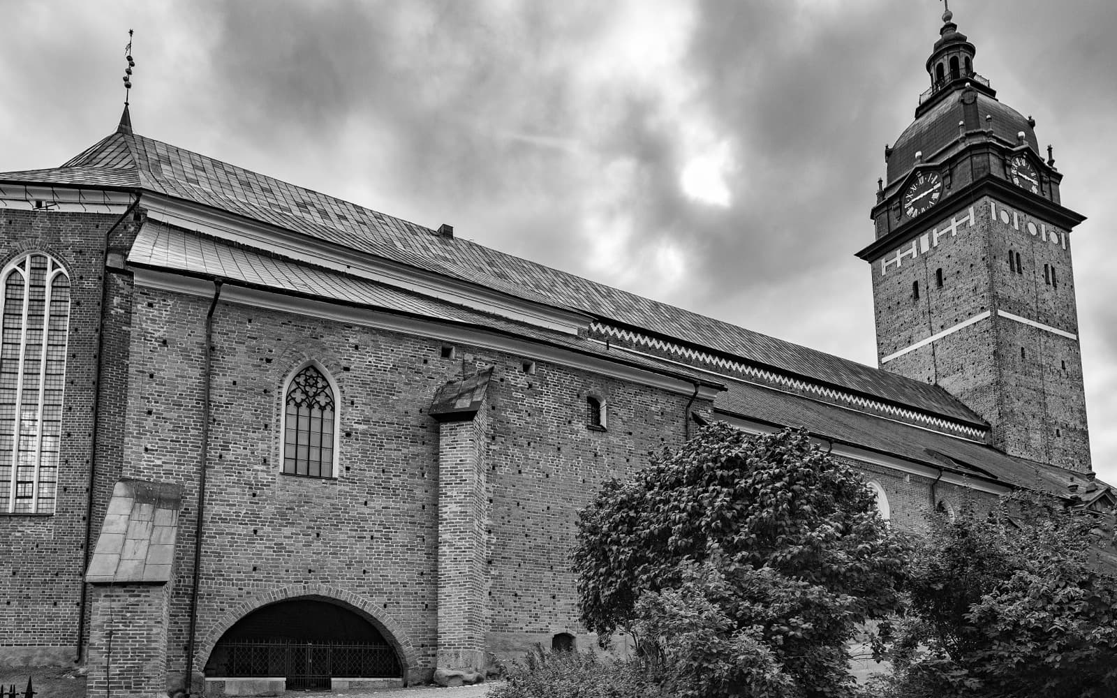 Svartvit bild av Strängnäs domkyrkas tegelfasad och klocktorn under mörka moln.