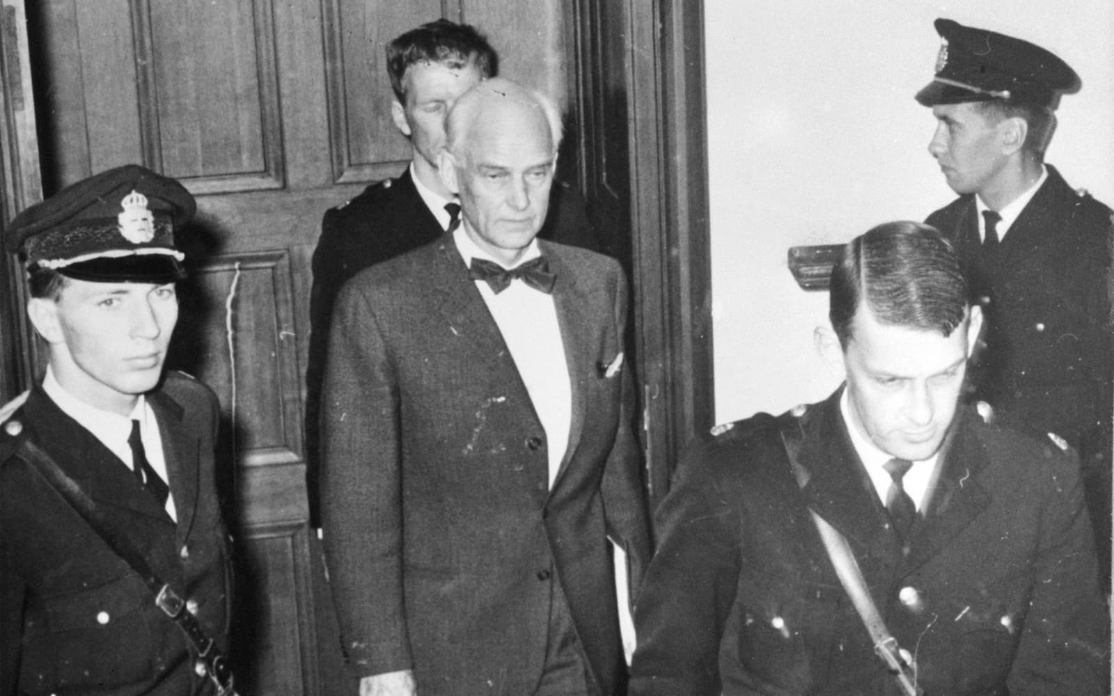 Svartvitt foto av en äldre man i fluga som leds ut genom en dörr av fyra poliser i uniform