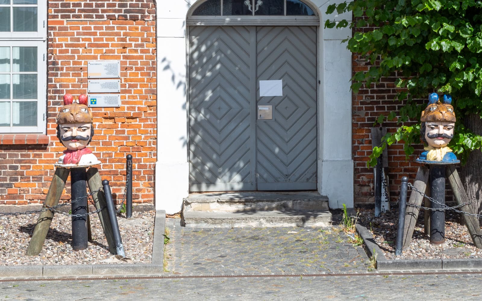 Två målade byster med lejonhuvuden som mössor står på stolpar framför en tegelbyggnad i Wismars hamn.